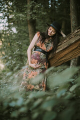 Fototapeta na wymiar Pregnant woman posing in the autumn forest. Portrait of a happy and pretty pregnant woman looking at her belly, leaning on the tree outdoors