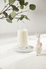 A glass of milk on a marble plate on a white table cloth, freshly baked cookies, branch of green eucalyptus , and white figurine of an easter bunny