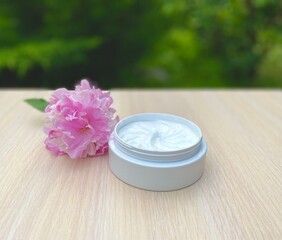 White skin face cream for body and hands for cosmetic spa treatments in a white jar next to a pink peony flower on a wooden table against a background of green plants outdoors.