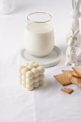 A glass of milk on a marble plate on a white table cloth, freshly baked cookies, creamy bubble candle and white easter bunnies, figurines