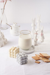 A glass of milk on a marble plate on a white table cloth, freshly baked cookies, bubble candles and white easter bunnies, figurines