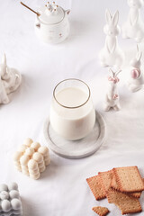 A glass of milk on a marble plate on a white table cloth, freshly baked cookies, bubble candles and white easter bunnies, figurines