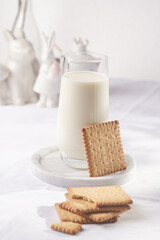 A glass of milk on a marble plate on a white table cloth, freshly baked cookies, and white easter bunnies, figurines