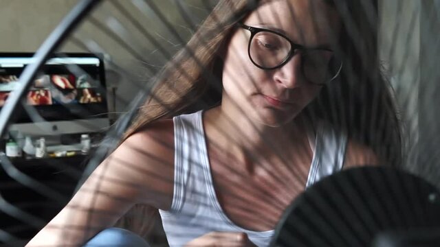 Tired overheated young hipster woman wear glasses suffering from too high temperature in home office sitting in front cooling fan, ventilator. Hot weather. People texting phone check social media.