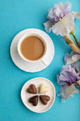 Obraz na płótnie Canvas Cup of cioffee with chocolate candies and lilac iris flowers on blue pastel background. top view, close up.