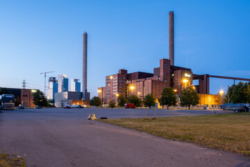 An old power plant with two smoke stacks. Modern skyscrapers in the background.