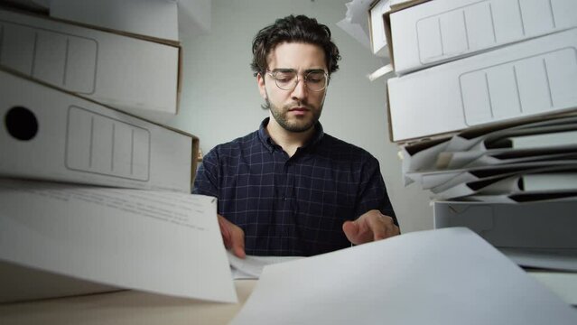 Tired dark-haired man with pile of folders working in office, takes glasses off, employee works with documents having headache, exhausted worker holding his head, red eyes, fatigue, lots of work. 