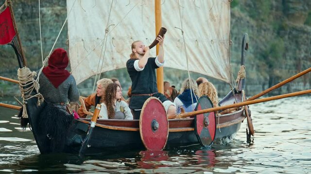 Vikings Sail on an Old Ship with a Raised Sail on a Calm River Against the Backdrop of a Rocky Coast. The Man Blows the Horn. Medieval Reconstruction.
