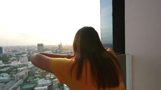 Beautiful Asian woman standing on apartment balcony looking away to cityscape and skyscraper at sunset with happiness. Smiling female relax and enjoy city life and indoor lifestyle at condominium