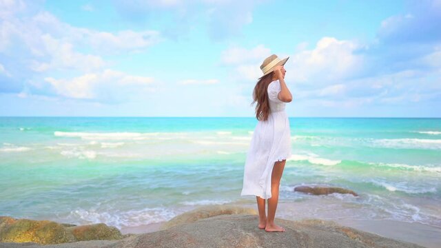 Lonely sexy exotic female in a white dress standing on rock in front of tropical sea waves holding floppy summer hat, full frame slow motion