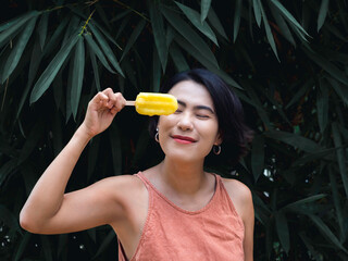 Woman eating popsicles. Happy beautiful Asian woman wearing casual pink tank top holding yellow popsicle on green tropical palm leaves background, outdoor. Smiling female enjoying ice lolly in summer.