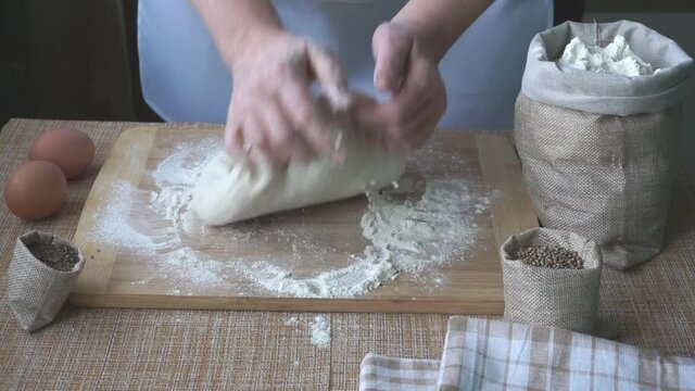The hands of an elderly woman knead the dough for bread on a wooden board on the table, on the table there are 2 eggs, there are linen bags with spices cumin and coriander and flour. World Bread Day