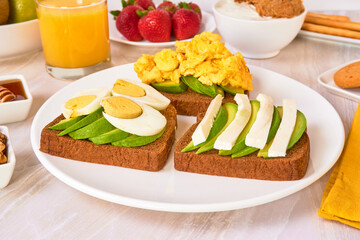 3 toasted breads with avocado, hard boiled egg and scrambled eggs on a white plate, accompanied by Greek yogurt and fresh fruit on a marble surface and white background. close up