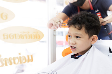 Cute asian child boy getting hair cut by hairdresser at hair salon. Hair care and childhood lifestyle concept.