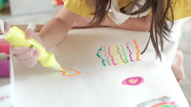Asian girl in yellow shirt and apron is drawing hearts shaped with multi-color glitter paint on white paper at home. Art class makes children creative. Playful activities to improve school skills.