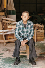 Portrait of tired pensive experienced senior carpenter sitting on stool he made at workshop resting after finishing work on big project