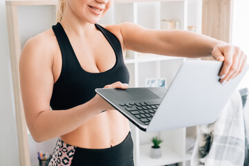Fitness trainer. Slim woman. Online sport. Home gym. Wellness body. Unrecognizable happy athletic lady in black sportswear opening laptop cover in hands light room interior.