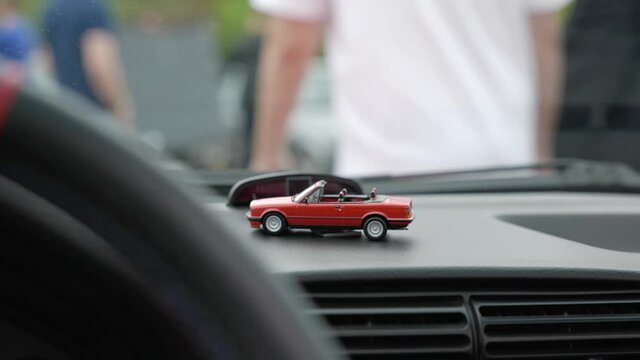 Tiny red toy car displayed on vehicle dashboard interior as people walk outside