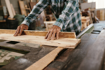 Hands of senior carpenter in plaid shirt cutting wide pine plank with circular saw