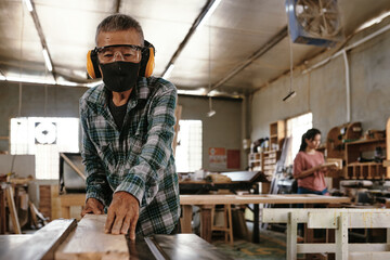 Senior carpenter in protective face mask and earmuffs cutting long wooden plank on workbench