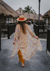 Naklejka premium Back view of a girl with beautiful long hair wearing an orange dress and a straw hat walking gracefully in a summer luxury resort