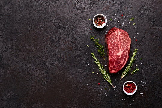 Fresh raw marbled beef rib eye steak, herbs and spices on black stone background, copy space