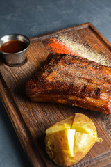 Grilled pork ribs with baked potato and sauce on a wooden board over stone grey background.