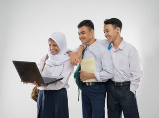 Three teenagers in junior high school uniforms smiling using a laptop computer together while carrying a backpack and a book on an isolated background