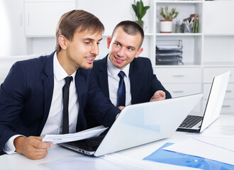 Two positive business men colleagues working together with laptops on desk in office