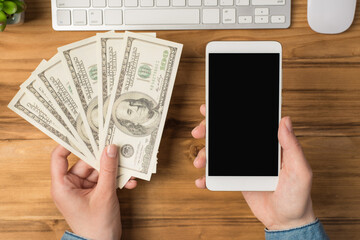 First person top view photo of female hands holding fan of money hundred dollars banknotes and smartphone flowerpot white keyboard and mouse on isolated wooden table background with copyspace