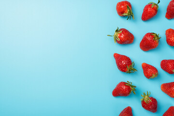Top view photo of scattered strawberries with leaves on the right on isolated pastel blue background with copyspace on the left