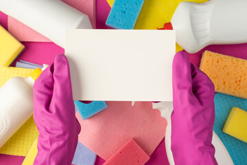 Top view photo of hands in pink rubber gloves holding white paper card detergent bottles multicolor napkins scouring pads on isolated pink background with copyspace