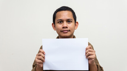 A portrait of young Asian Malay man with casual brown shirt holding an empty blank paper on isolated white backgrounds. Empty space for text.