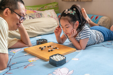 Asian cute child girl playing Go or Chinese traditional board game on bed, The girl is thinking, and father waiting for her move.