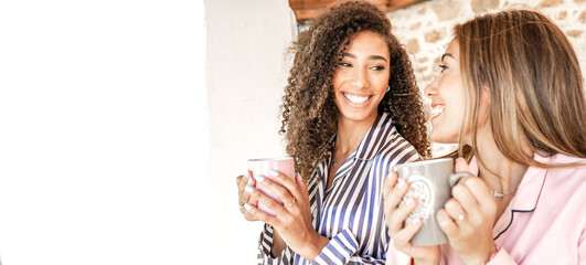 Multiracial women couple smiling looking each other holding a teacup in pajama just waken up. New...