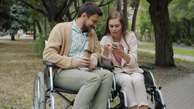 Disabled couple in wheelchairs are using smartphone drinking to go coffee and talking outdoors in city park. Relationship and leisure time concept.