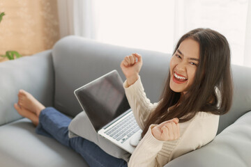 Young happy attractive Asian woman wearing casual clothes using laptop computer and relax sitting on couch sofa at home. Excited women rising hands with success or close the deal. Online job concept.