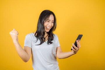 Fototapeta premium Portrait of happy Asian woman wears white t-shirt raise hands glad excited cheerful and holding mobile phone or smartphone isolated on bright yellow background