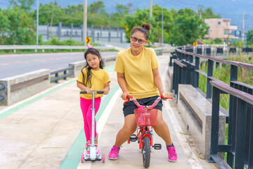 Little child girl to ride scooter with mother ride on small bicycle  outdoor sports ground on sunny summer day. Active leisure and outdoor sport for children.