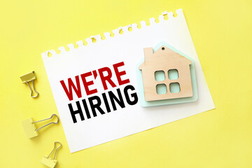 We're Hiring . text on white paper on yellow background. with a picture of a wooden house