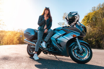 Motorcycle local travel. Young smiling woman in a leather jacket and sunglasses sitting on a motorbike and using a smartphone. World Motorcyclist Day concept