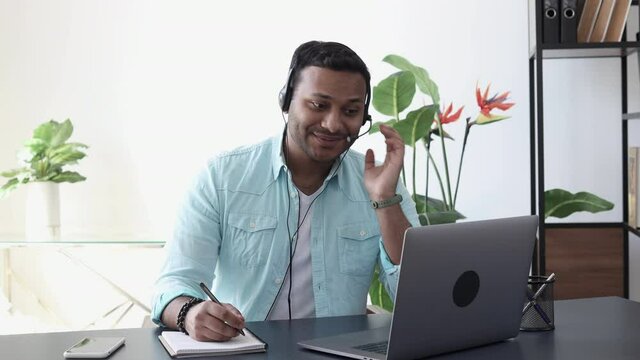 Young indian man in headset with headphones and laptop at the table writes important information in a notebook. Talking, meeting online or e-education, support services