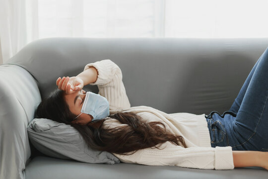 Young Asian woman wearing protective face mask stay quarantine at her apartment. Sick women coughing. Lonely lady catch a cold suffering lying on a couch at home
