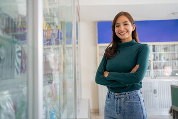 beautiful asian woman smiling with crossed hands while standing near cell phone accessories display case in cell phone shop