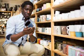 Focused african american man visiting household goods store in search of dishware, looking for new cups