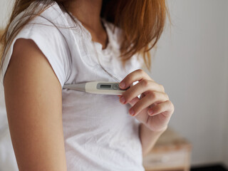 A woman in a white t-shirt is checking the temperature with a thermometer under her arm