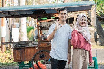 proud couple while selling food on the street. young muslim small business entrepreneur with their food cart at the background showing thumb up