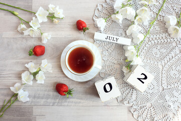 Calendar for July 2 : cubes with the numbers 0 and 2, the name of the month of July in English, a cup of tea on a gray openwork napkin, scattered flowers of bluebells and strawberries, top view