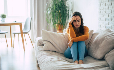  Young sad Brunette woman with severe headache holds her head while she sitting on the couch at home