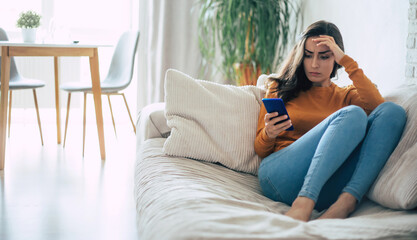 Fototapeta premium Frustrated and depressed young brunette woman is crying with a smartphone in hands while she sitting on the couch at home